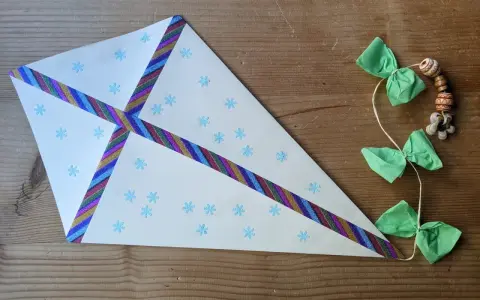 Bastelanleitung mit Bildern: Ein selbstgebastelter Drachen aus Papier mit Schneeflocken und buntem Klebeband, geschmückt mit grünen Schleifen und Holzperlen.