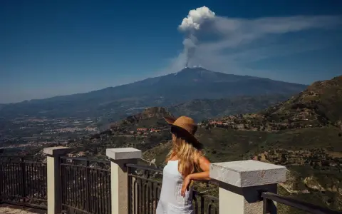Frau mit Hut blickt auf den rauchenden sizilien vulkan. Die Landschaft ist grün und bergig.