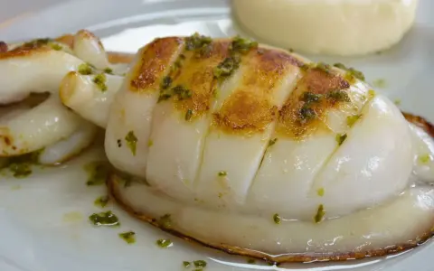 Delicioso sepia a la plancha con hierbas verdes, servido en un plato blanco con una guarnición cremosa.