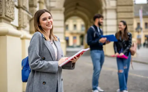 Uśmiechnięta studentka z notatnikiem w ręku, przygotowująca się do studiów 2 stopnia. W tle para studentów.