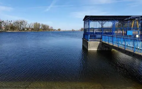 Spokojne jezioro średzkie z błękitną budowlą i pomostem. W tle drzewa i zabudowania.