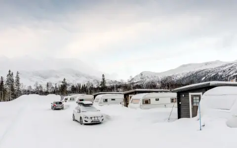 Vinterlandskap vid moskosel camping med snötäckta husvagnar och bilar, berg i bakgrunden.