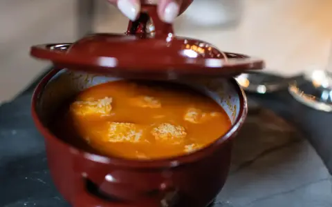 Una mano levanta la tapa de una cazuela de barro con una deliciosa comida típica de Ávila, un guiso naranja con trozos blancos.