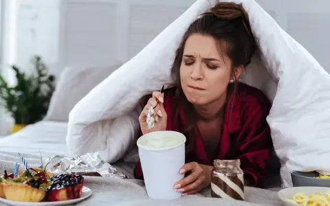 Ragazza sotto le coperte mangia gelato, dolci e pasta. La sua espressione suggerisce una forte fame pre ciclo.