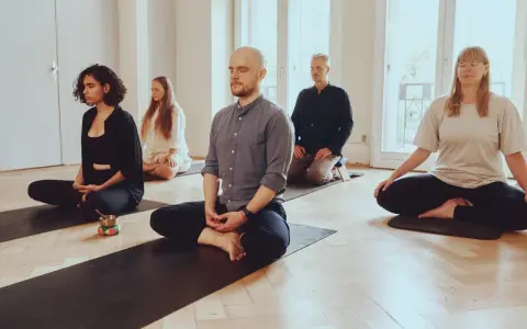 Menschen in einem hellen Raum meditieren auf Matten. Die Meditation wirkt beruhigend und fördert die Konzentration.