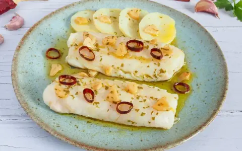 Dos filetes de merluza a la bilbaina, con patatas, ajo laminado y guindilla, en un plato azul.