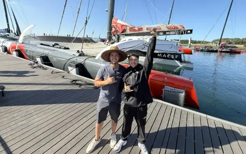 Deux jeunes hommes posent devant un bateau de course prêt pour une traversée atlantique. Le bateau est long, gris et rouge, avec des voiles partiellement visibles.