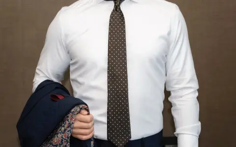 Elegancki mężczyzna w białej koszuli i modnym krawacie męskim w kropki, trzymający granatową marynarkę z wzorzystą podszewką.
