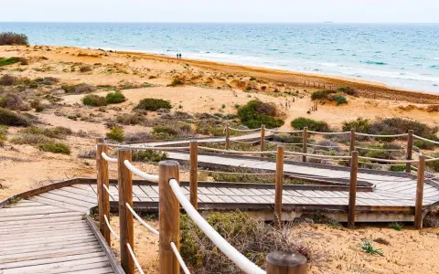 Pasarela de madera serpentea por la duna, invitando a explorar la ruta Calblanque hacia el mar azul.