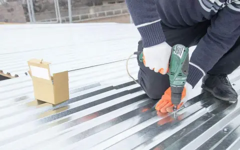 Pose sur une toiture bac acier isolé, un ouvrier visse une plaque avec une perceuse. Une boîte de vis est à proximité.