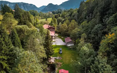 Idyllisches **Camping in den Bergen**: Ein Wohnmobil steht auf einer grünen Wiese neben einem Bach, umgeben von dichten Wäldern und traditionellen Holzhäusern.