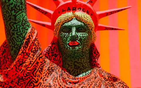 A vibrant Statue of Liberty sculpture, reimagined with Keith Haring's iconic political art style, features a green face with bold black lines and red lips, and an orange robe adorned with his signature figures.