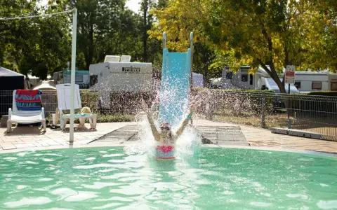 Radość na aranypart camping! Dziecko zjeżdża ze zjeżdżalni prosto do basenu, tworząc fontannę wody.