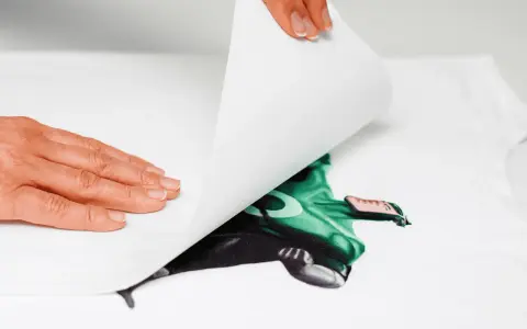 Manos aplicando una hoja para imprimir transfer sobre una camiseta blanca con un diseño de superhéroe verde.