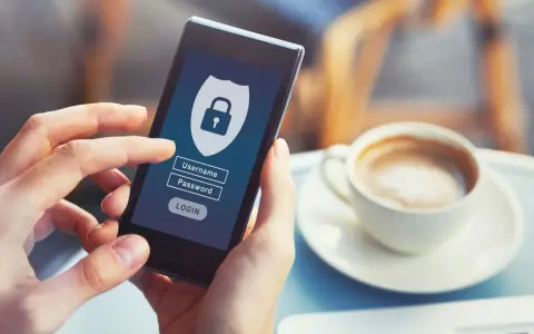 Dłoń trzyma smartfon z ekranem logowania do banku. Widoczny symbol tarczy z kłódką, pola na login i hasło.