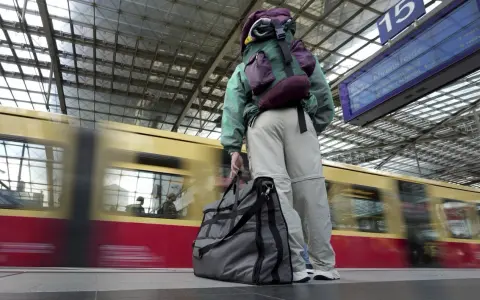 Traveler with backpack and duffel bag waits on a platform as a train speeds by.