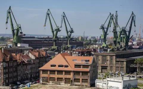 Widok na stocznię w Gdańsku z charakterystycznymi dźwigami. W oddali budynek z napisem "Nowe Syrie", nawiązujący do muzeum sztuki współczesnej Gdańsk.