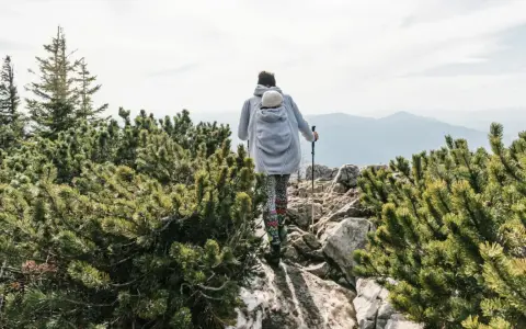 Frau wandert mit Baby im Tragetuch auf einem felsigen Bergpfad, umgeben von grünen Kiefern.
