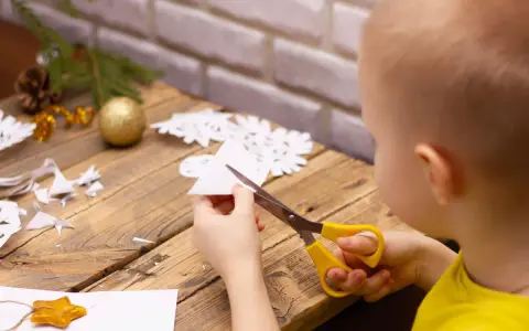 Małe rączki wycinają śnieżynkę z papieru. Na stole leżą gotowe ozdoby i gałązka choinki.