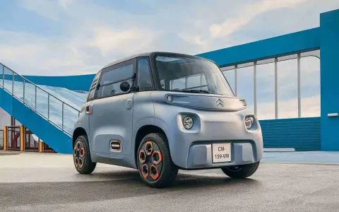 Un cuadriciclo pesado Citroën Ami gris con ruedas naranjas, aparcado frente a una estructura azul y una rampa.
