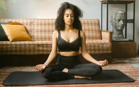 Frau in schwarzer Sportkleidung praktiziert Meditation für tiefe Entspannung auf einer Matte vor einem karierten Sofa.