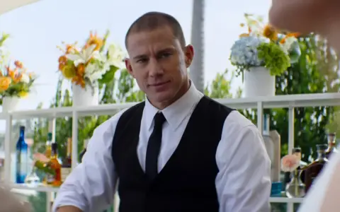 Channing Tatum som bartender i en film. Han ser ut att vara redo att servera drinkar med blommor och flaskor i bakgrunden.