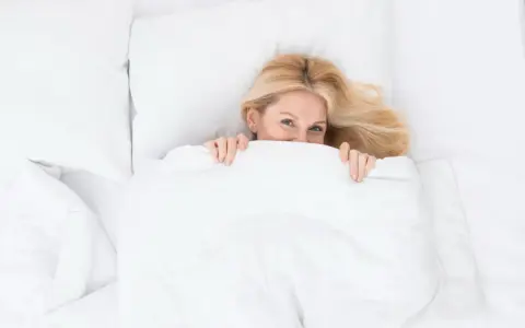Uśmiechnięta blondynka ukrywa się pod białą kołdrą. Czy to znak, że zastanawia się, jak prać kołdrę?