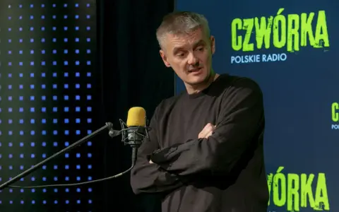 Jacek Sienkiewicz w studiu Polskiego Radia, przed mikrofonem, z rękami skrzyżowanymi na piersi.