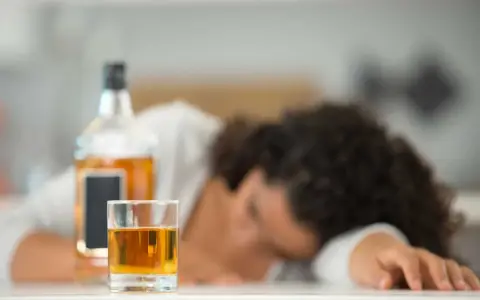 Szklanka z alkoholem i butelka na stole, osoba śpiąca w tle.