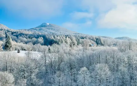 Bieszczady zimą. Ośnieżone drzewa i góry pod błękitnym niebem. W oddali widać mały domek.