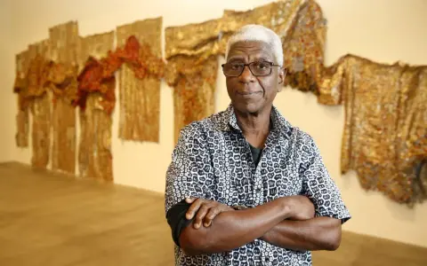 A distinguished man with white hair and glasses, arms crossed, stands before a wall adorned with shimmering, textured artworks, a testament to the enduring legacy of famous artists today.