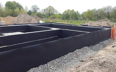 Fundament domu w trakcie budowy, z czarną membraną i otoczakami. Trwa ocieplenie fundamentów, przygotowanie do dalszych prac.