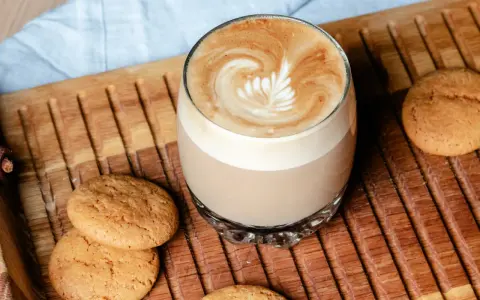 Ein Glas Flat White mit Latte Art neben Keksen auf einem Holztablett. Der Unterschied zwischen Flat White und Cappuccino ist hier deutlich sichtbar.