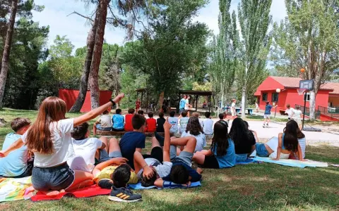 Niños disfrutando de actividades al aire libre en campamentos de verano aula joven. Un grupo observa un partido de baloncesto mientras otros descansan en el césped.