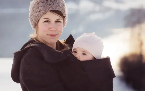 Kvinna med bebis i bärsele, båda med mössor på. Vinterlandskap i bakgrunden.