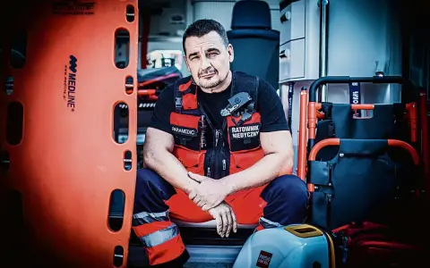 Ratownik medyczny w kamizelce z napisem "Paramedic" i "Ratownik Medyczny" siedzi w karetce obok noszy.
