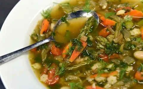 Un fumante minestrone di verdure, ricco di carote, fagioli e erbe aromatiche, servito in una ciotola bianca con un cucchiaio.