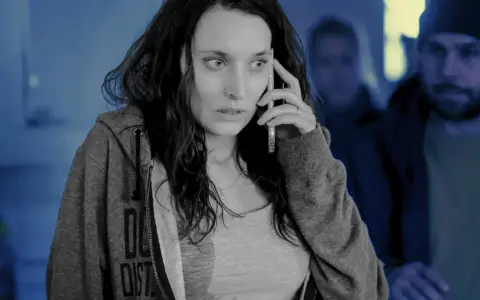 Przerażona kobieta rozmawia przez telefon, podczas gdy w tle widać zamazane postacie. Scena jak ze skandynawskich seriali kryminalnych.