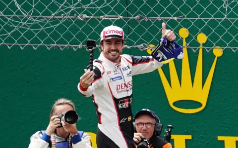 Fernando Alonso celebra su victoria en Le Mans, con una gorra y un trofeo. Fotógrafos capturan el momento.