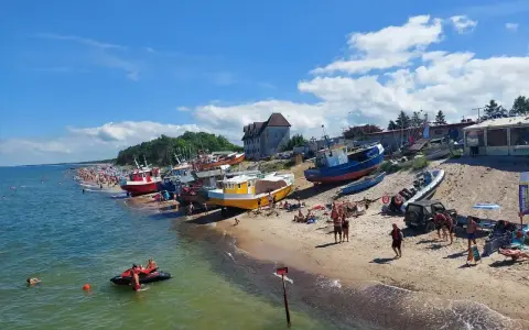 Słoneczny dzień na plaży, kolorowe łodzie na piasku, tłumy ludzi szukają idealnych miejsc na wakacje.