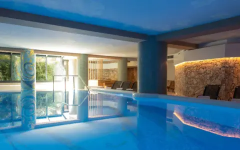 Indoor swimming pool with lounge chairs and a stone wall with warm lighting.