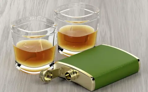 Zwei Gläser mit bernsteinfarbenem Whiskey und ein grüner Taschenflachmann. Ein 4 cl Whiskey hat ca. 90 Kalorien.