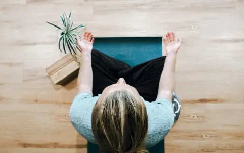 Person mediterar på en yogamatta, en grön växt och träblock bredvid. Perfekt för avkoppling på arbetsplatsen.