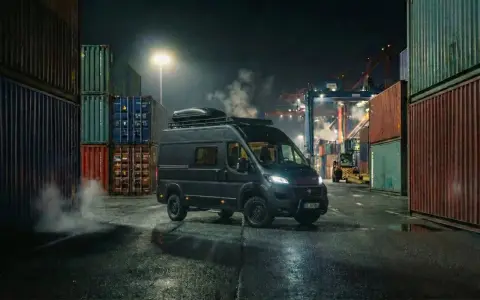 Mörk Fiat Ducato står vid en hamn, omgiven av containrar. Rök stiger från marken, kanske ett tecken på fiat ducato automatlåda problem.