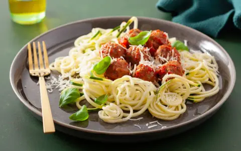 Pyszna keto kolacja: klopsiki w sosie pomidorowym na cukinii spaghetti, posypane parmezanem i listkami bazylii. Złoty widelec czeka.