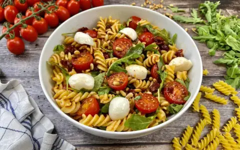Ein köstlicher Pesto-Salat mit getrockneten Tomaten, Mozzarella-Kugeln und Pinienkernen, serviert in einer weißen Schüssel.