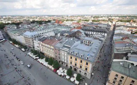 Widok z góry na Rynek Główny w Krakowie. Tłumy ludzi spacerują, a dorożki czekają na turystów. W tle widać zabytkowe kamienice i rozległą panoramę miasta.
