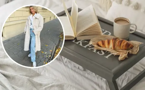 Kobieta w płaszczu z owczej wełny, symbolizującym quiet luxury, cieszy się śniadaniem w łóżku: książka, croissant i kawa.