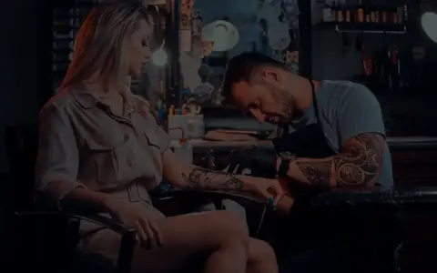 Artysta tworzy tatuaż opaska na nodze kobiety. W tle widać studio tatuażu.