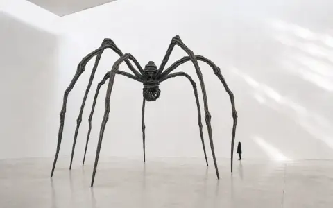 Une araignée monumentale, œuvre d'un artiste contemporain, domine une salle d'exposition. Une silhouette humaine, petite, contemple cette sculpture imposante.